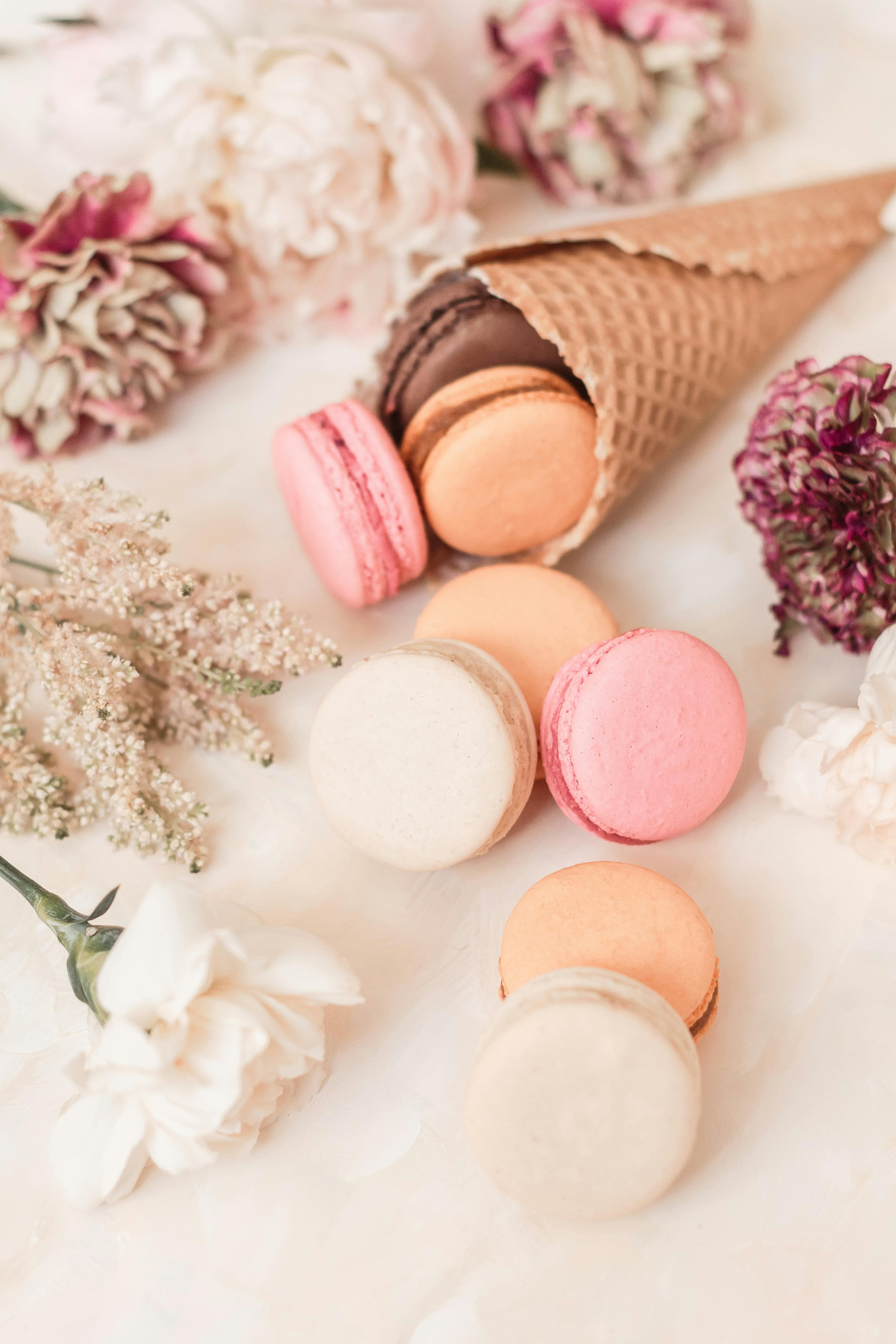 A delicate arrangement of colorful macarons spilling from a waffle cone, interspersed with blooming flowers, on a soft pastel backdrop.