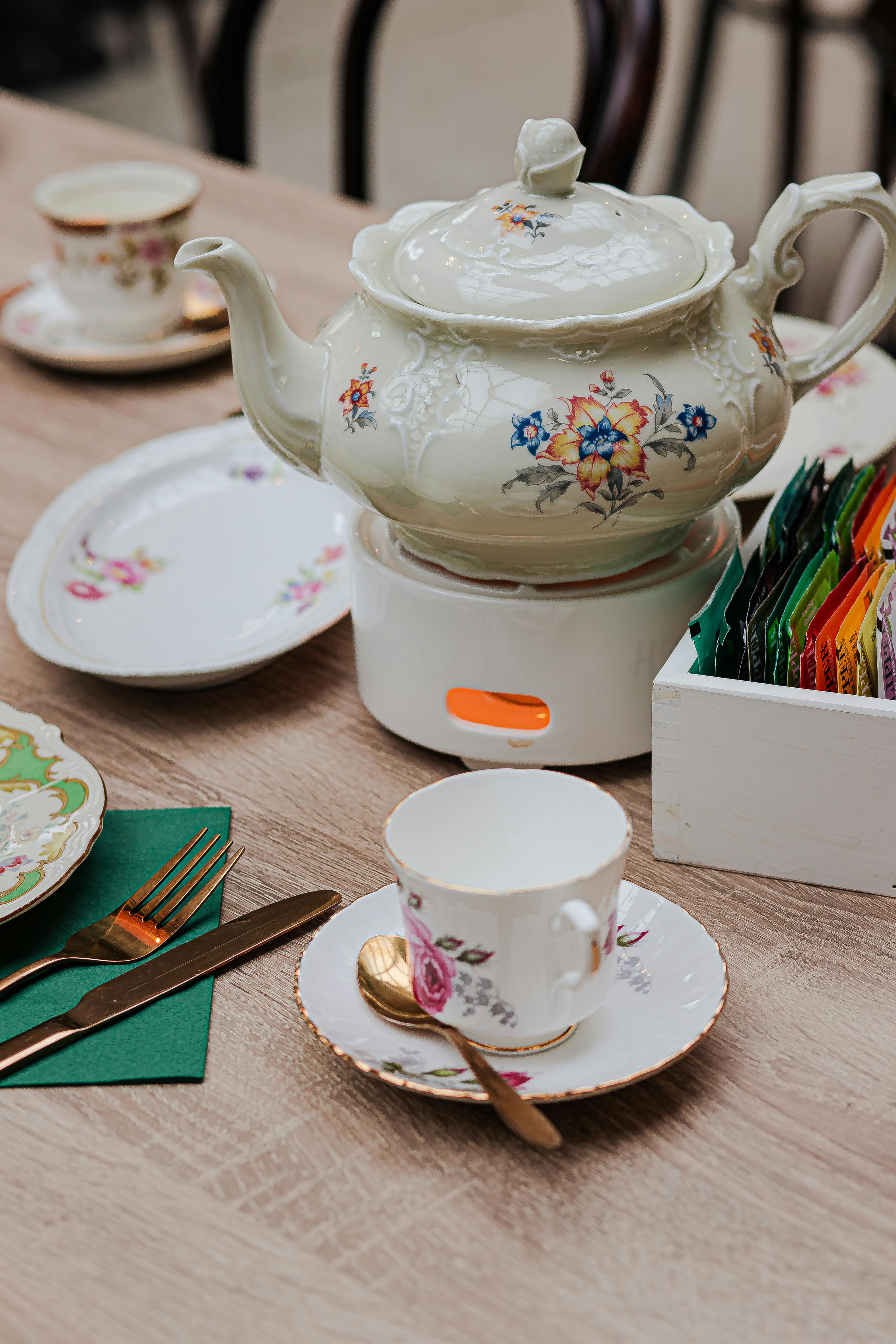 An elegant tea service arrangement featuring a floral-patterned teapot on a warmer, a matching teacup on a saucer with a gold spoon, plates with similar designs, green napkins, and gold cutlery, alongside a box of colorful tea packets.