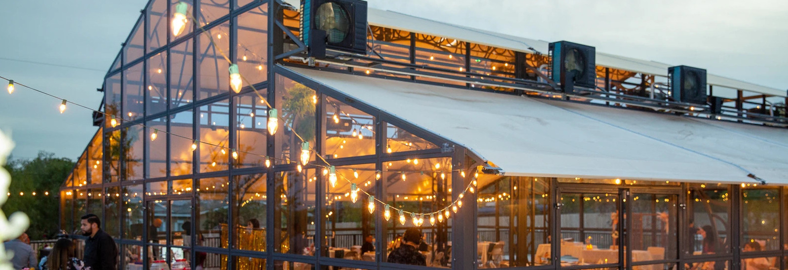 Dusk View of a Modern Glasshouse Event Venue Elegant glass greenhouse venue adorned with string lights at dusk, showcasing interior warm lighting and exterior spot cooling units.