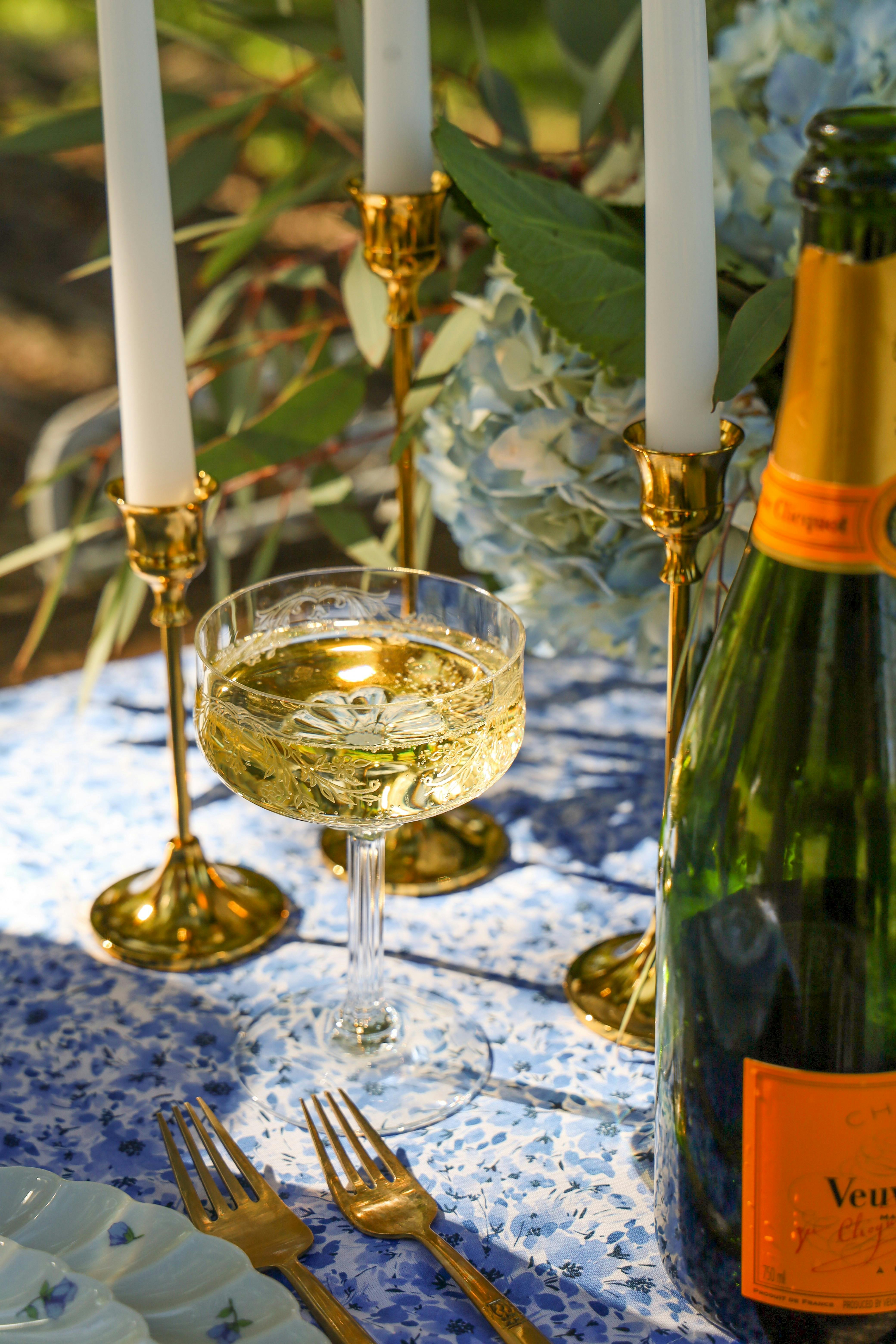 Elegant outdoor dining setup with a champagne glass and bottle, gold candlesticks with white candles, and gold cutlery on a blue floral tablecloth.