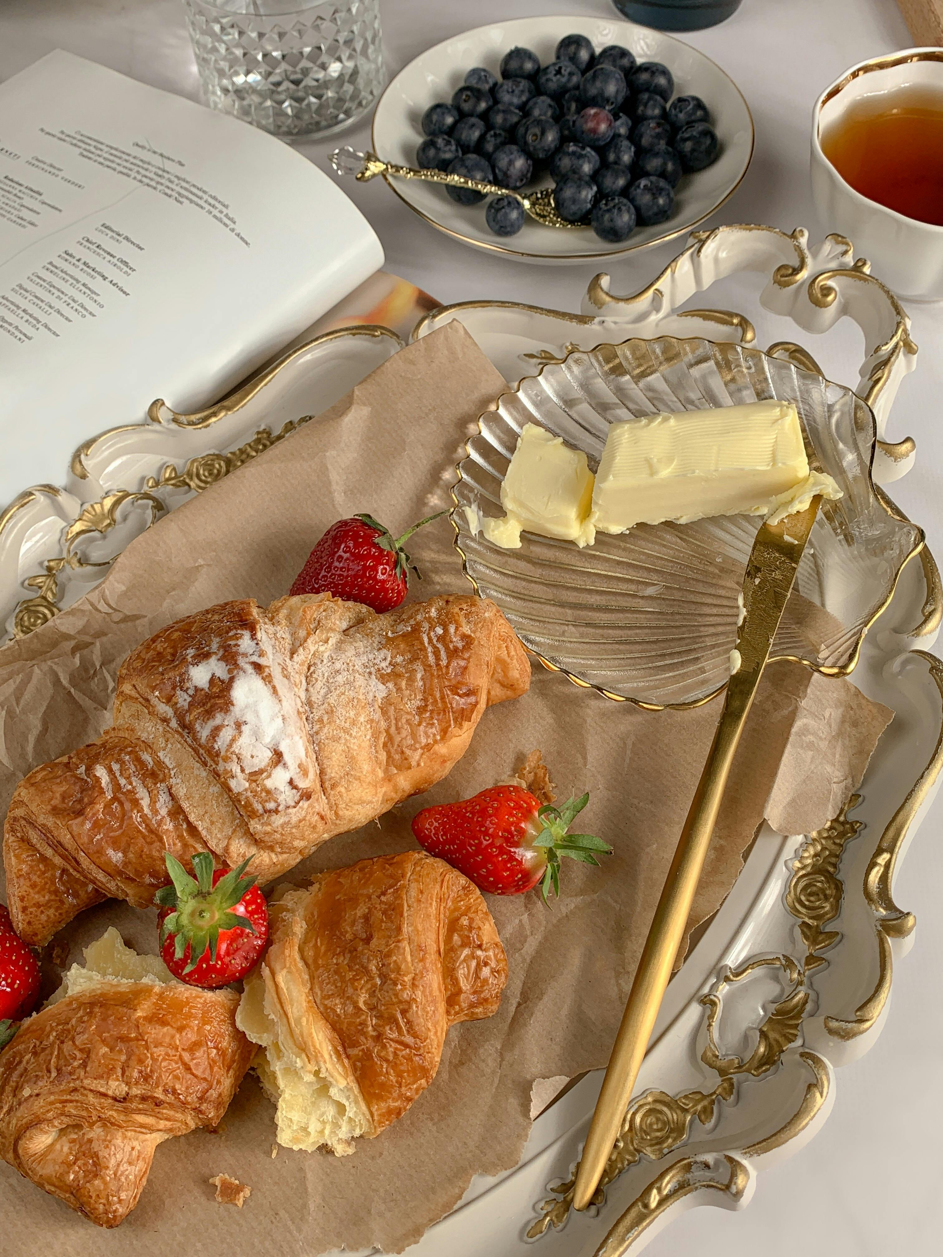 High tea setting with fresh croissants, strawberries, blueberries on a golden tray, butter on a glass dish with a gold knife, and a cup of tea with a menu in the background.